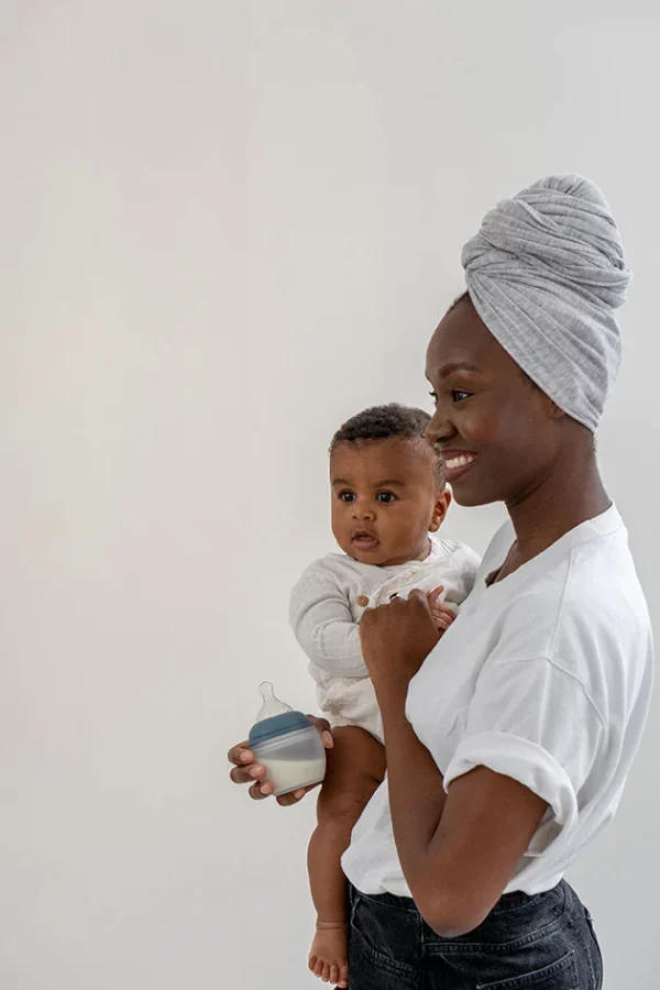 une maman portant son bebe en tenant dans une main un biberon élhée
